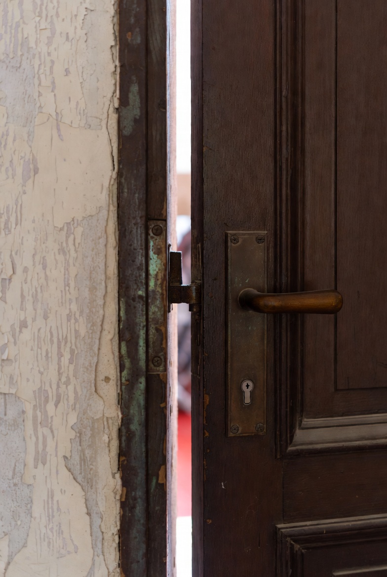 A photograph of an open wooden door with hinge and lock, revealing a dimly lit interior in a decayed setting.