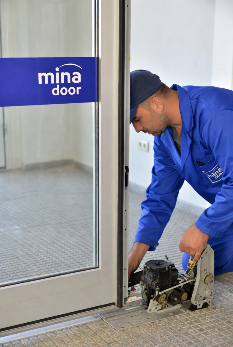 A technician repairs an automatic sliding glass door with "mina door" logo in a public space.