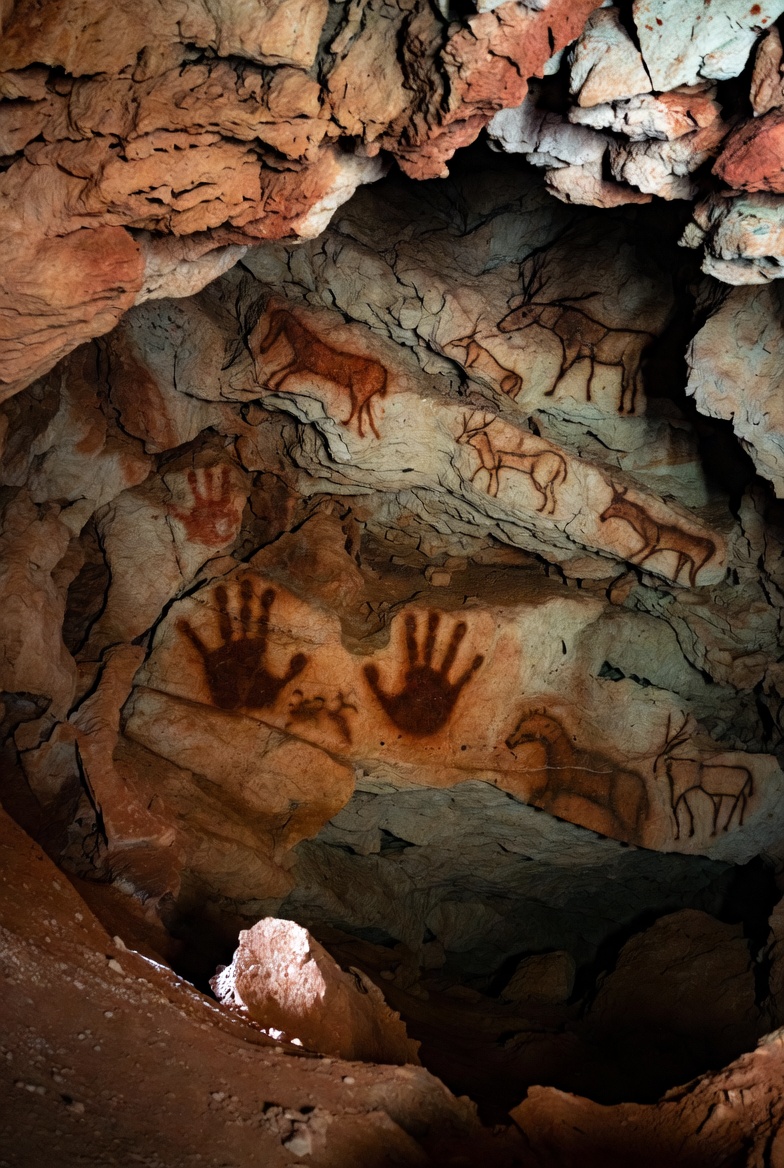 A photograph of ancient cave wall art with prehistoric handprints and animal drawings in reddish-brown hues.